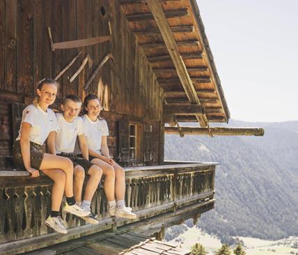 Three smiling teenagers in traditional clothing and white T-shirts sit on the wooden balcony of an alpine hut overlooking a sunny valley and forested mountain slopes