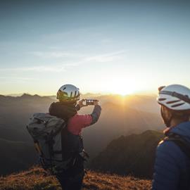 Due hikers are watching the sunrise from a mountain peak. One is taking a picture with his smartphone.