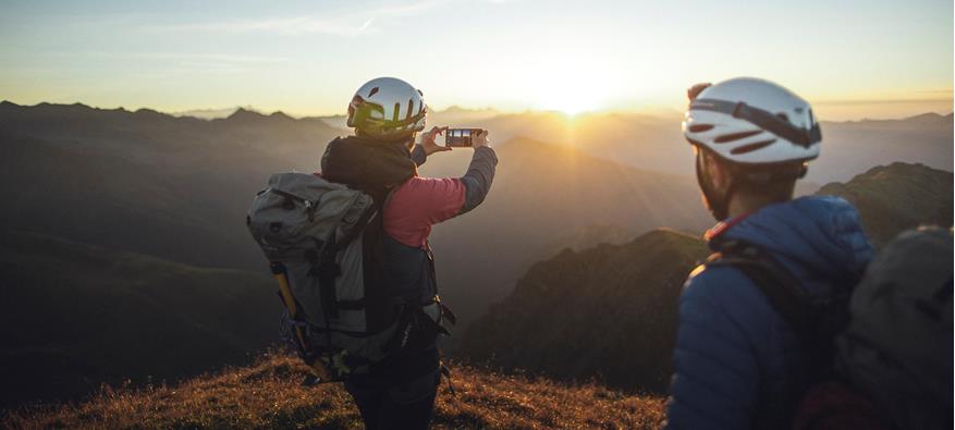 Zwei Bergsteiger beobachten den Sonnenaufgang von einem Gipfel. Einer der beiden macht ein Foto mit dem Smartphone.