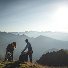 Two hikers are packing their backpacks at sunrise on a mountain ridge, surrounded by a vast alpine landscape with soft plays of light and shadow