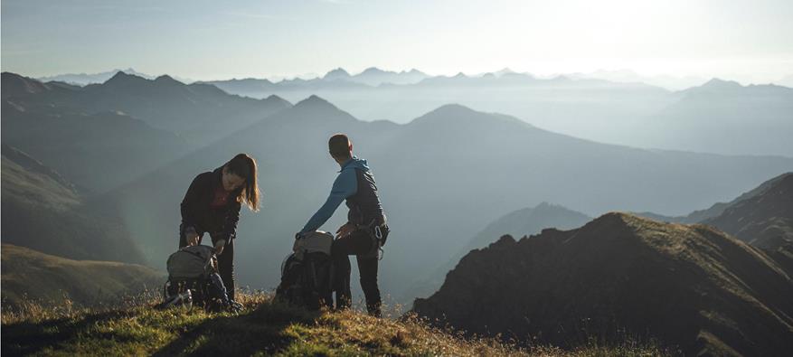 Due escursionisti sistemano i loro zaini all'alba su una cresta di montagna, circondati da un ampio panorama alpino con delicati giochi di luce e ombra.