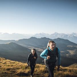 Two hikers are ascending through an alpine landscape under a clear blue sky, with striking mountain ranges stretching across the background