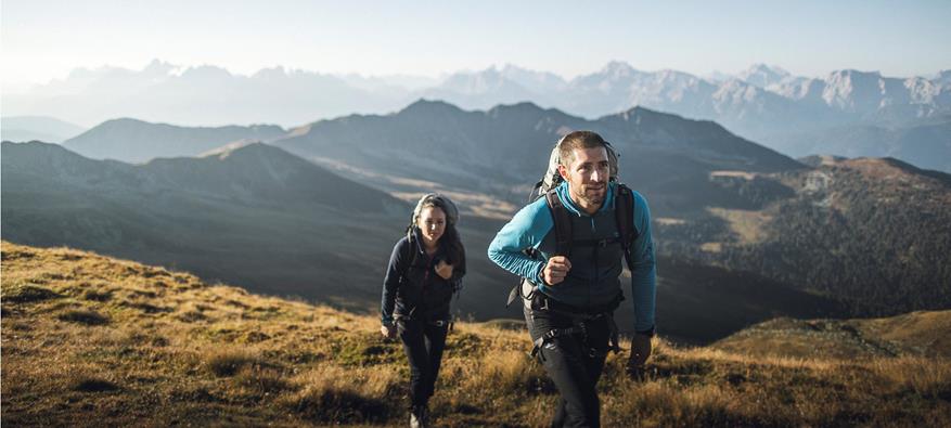 Two hikers are ascending through an alpine landscape under a clear blue sky, with striking mountain ranges stretching across the background