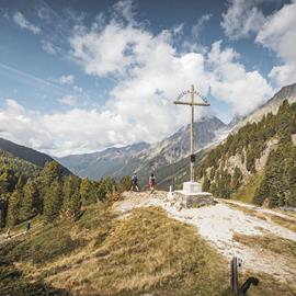 Two hikers at a wooden memorial cross inscribed ‘Unseren Helden’ on a scenic viewpoint surrounded by alpine mountains, forests, and open sky
