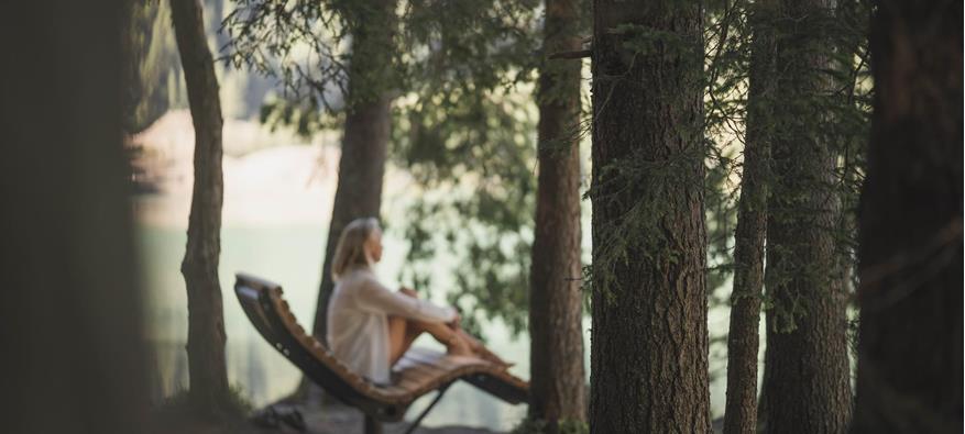 Una donna siede rilassata su una chaise longue in legno tra alti alberi al margine del bosco, guardando verso un lago.