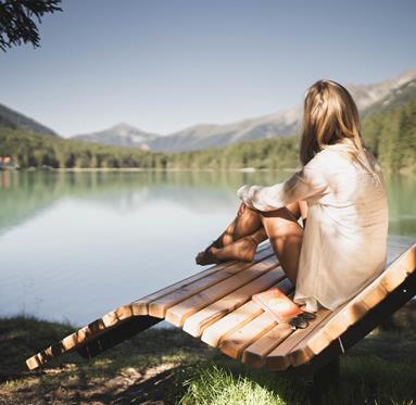 A woman sitting relaxed on a wooden bench by the lakeshore, gazing at the calm water and surrounding mountains; a book (The Alchemist) and sunglasses lie on the bench.