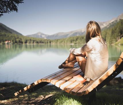 Eine Frau sitzt entspannt auf einer Holzliege am Seeufer, blickt auf das stille Wasser und die umliegenden Berge; auf der Bank liegen ein Buch (The Alchemist) und eine Sonnenbrille