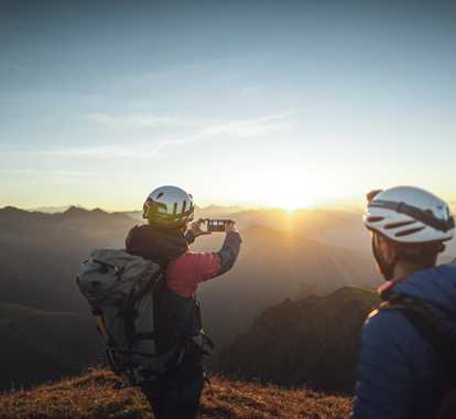 Zwei Bergsteiger beobachten den Sonnenaufgang von einem Gipfel. Einer der beiden macht ein Foto mit dem Smartphone.