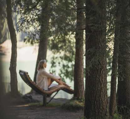 Una donna siede rilassata su una chaise longue in legno tra alti alberi al margine del bosco, guardando verso un lago.